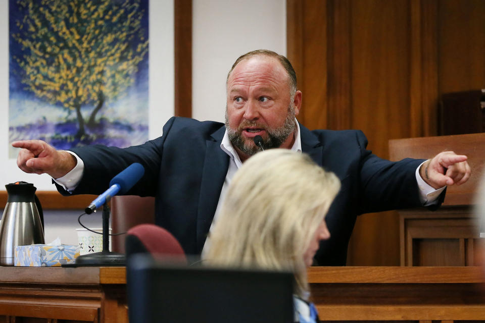 Alex Jones, sitting at a microphone, points during his testimony. 