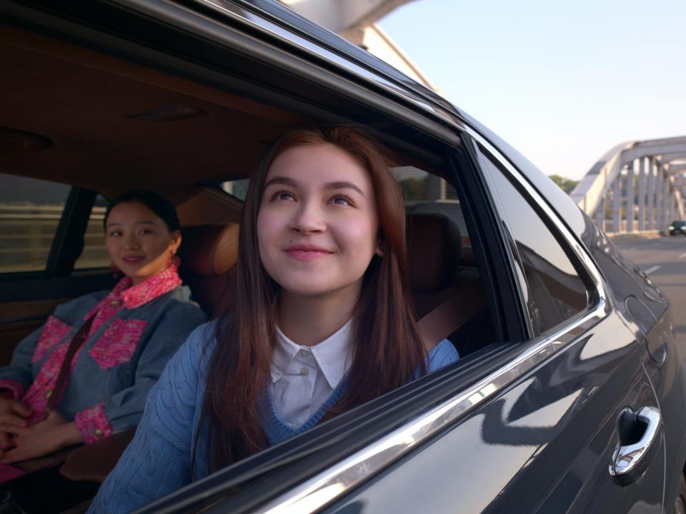 anna cathcart as kitty, staring wistfully out the window of a black car as it crosses a bridge. gia kim, as yuri, is visible in the background, looking at kitty