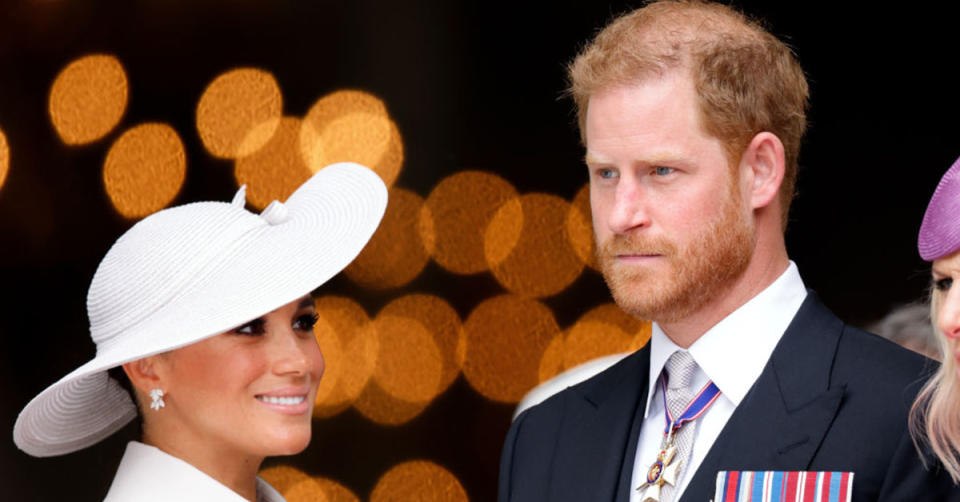 Meghan Markle wears a large white hat and smiles, next to her solemn husband Prince Harry wearing a suit jacket with medals