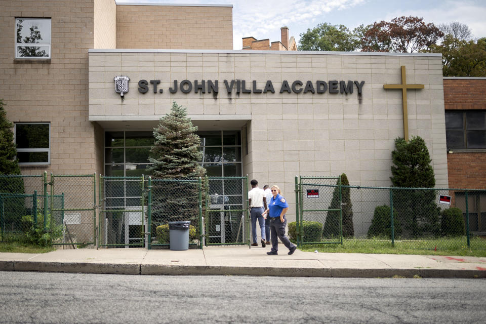 An NYPD community affairs officer escorts two individuals into the former Saint John Villa Academy being repurposed as a shelter for homeless migrants, Wednesday, Sept. 13, 2023, in the Staten Island borough of New York. Scott Herkert, a New Yorker upset that the city has been housing homeless migrants on his suburban block, has set up a loudspeaker to deliver an unwelcoming message in six languages to his new neighbors: "The community wants you to go back to New York City. Immigrants are not safe here." (AP Photo/John Minchillo)