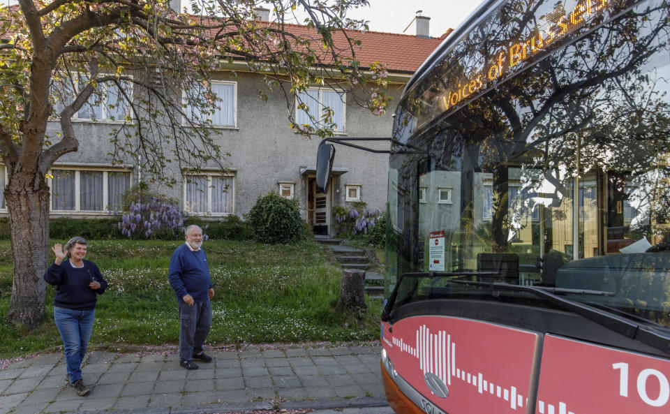 A couple come out onto the sidewalk to greet a bus delivering a loudspeaker message from family and friends in Brussels, Wednesday, April 22, 2020. With streets in the Brussels capital mostly devoid of loud traffic and honking horns, a simple piece of emotional poetry can split the air. The public bus company, STIB-MIVB, has been calling on people to send in voice messages, which are now delivered by a special bus driving in a loop to connect all the messages and leave a trail of happiness. (AP Photo/Olivier Matthys)