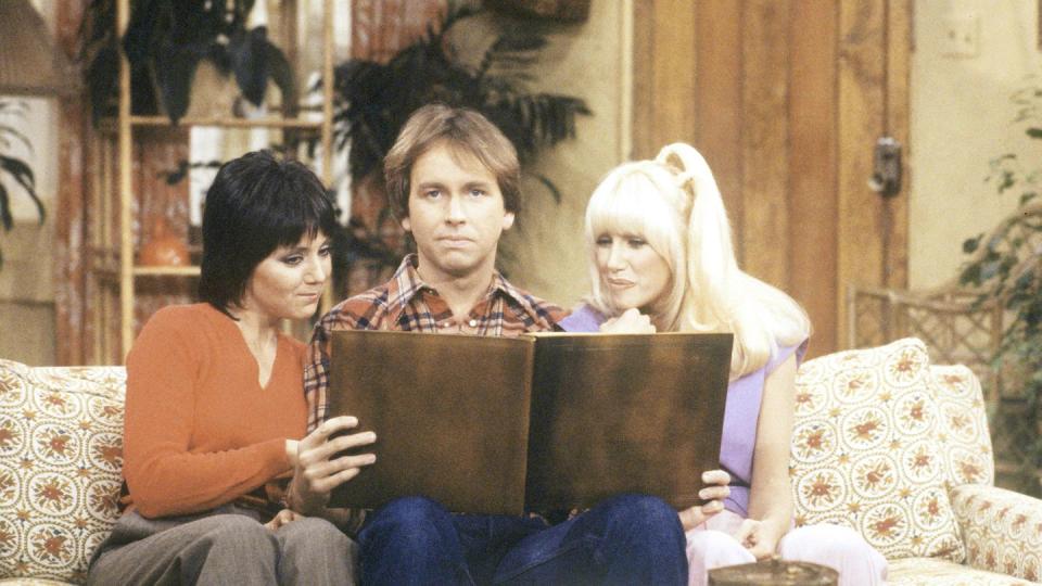 joyce dewitt as janet, john ritter as jack, and suzanne somers as chrissy in a scene from threes company, they sit on a couch in a living room and look at a book