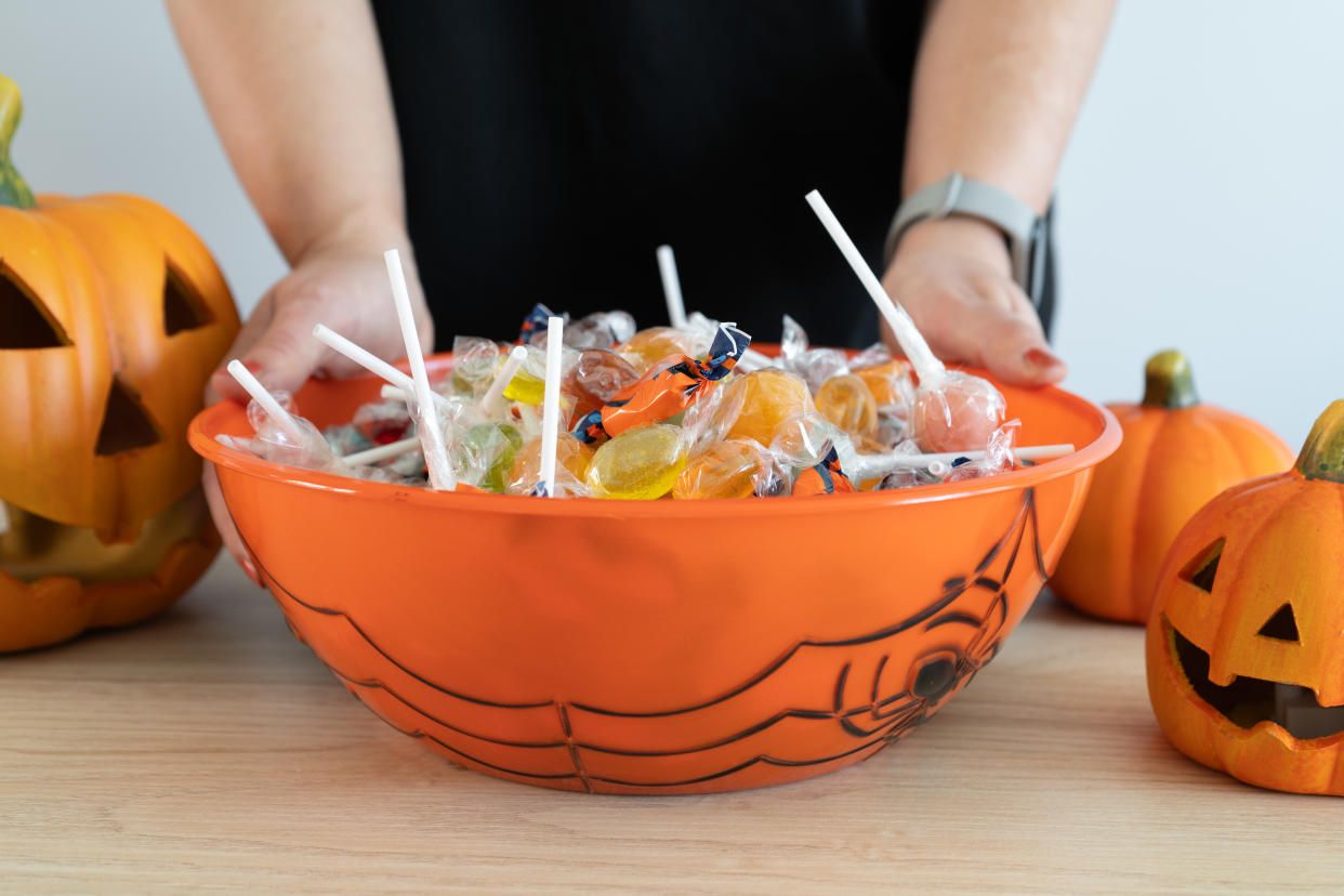 Woman holding a Halloween candy bowl filled with sweets for trick-or-treating. Female hands and Halloween-themed decorative treat bowl container. Jack-o'-lantern pumpkins placed around the table.