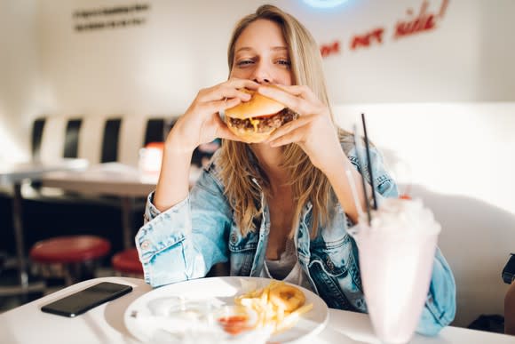 A woman sitting at a restaurant table eating a hamburger, french fries, and a milkshake.