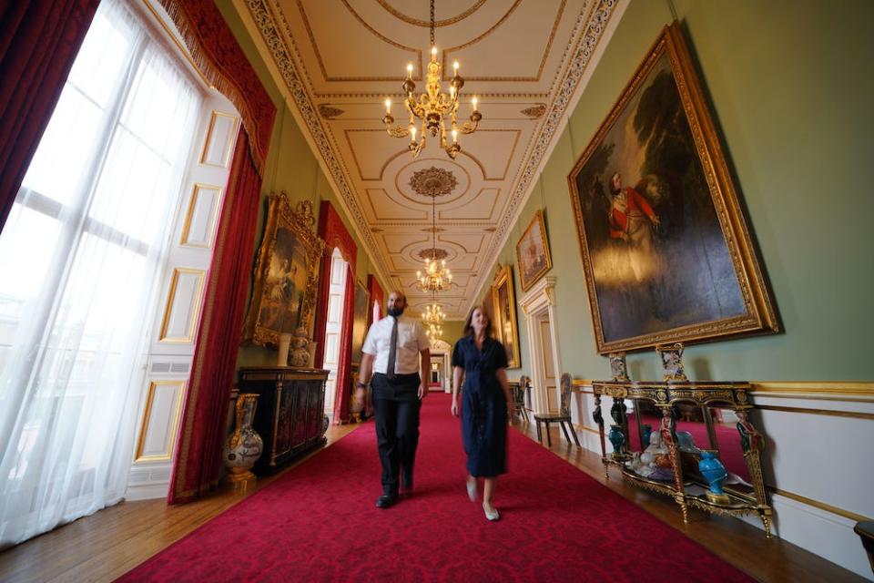Two Members of Royal Collection Trust staff walk down a long, red-carpeted corridor adorned with old paintings and ornate furniture and fixings on July 8, 2024, ahead of the opening of Buckingham Palace's East Wing following a long refurbishment.