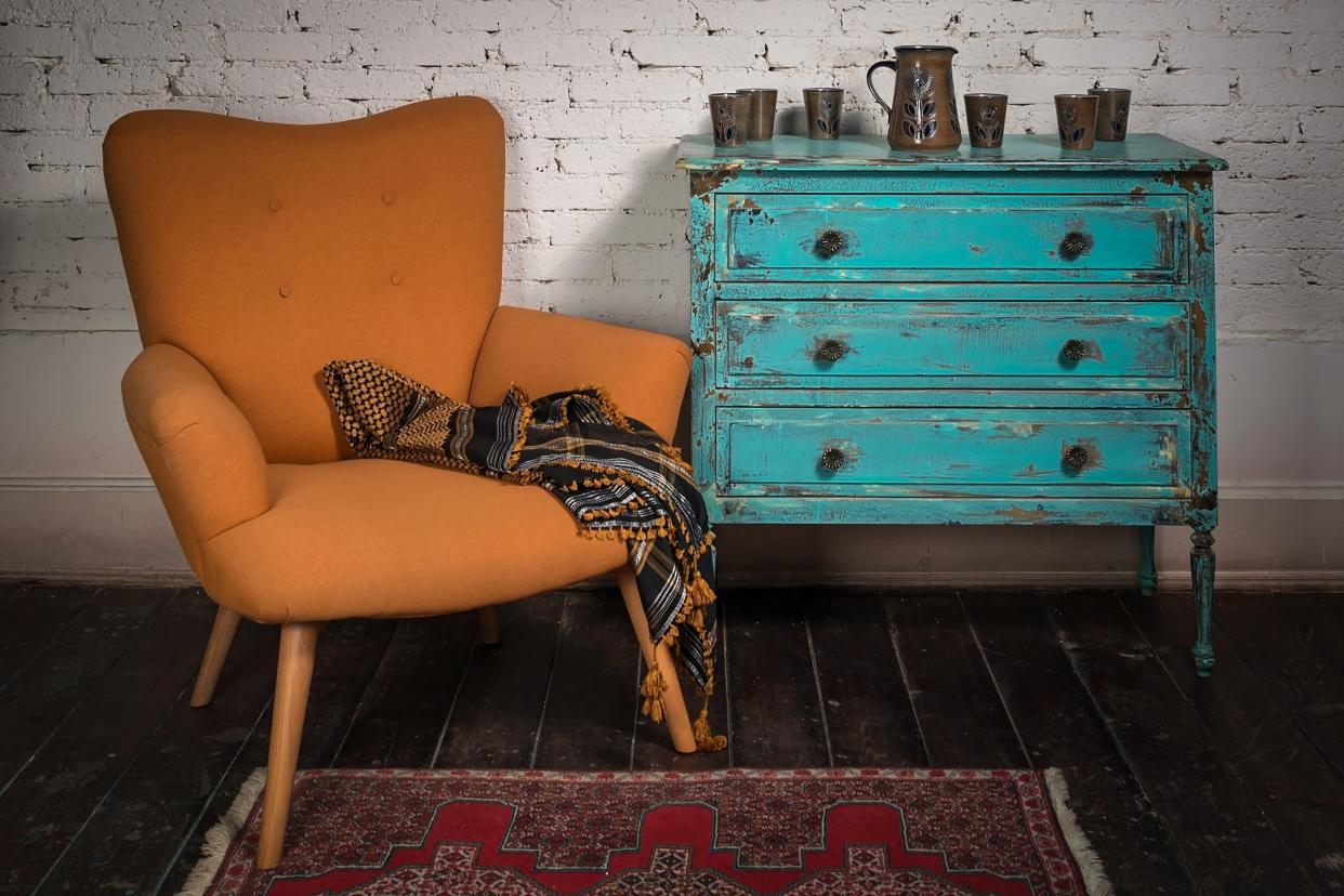 Ornate scarf on a vintage orange armchair on a maroon rug with a vintage blue cabinet on a worn dark brown wooden floor with a white painted brick wall