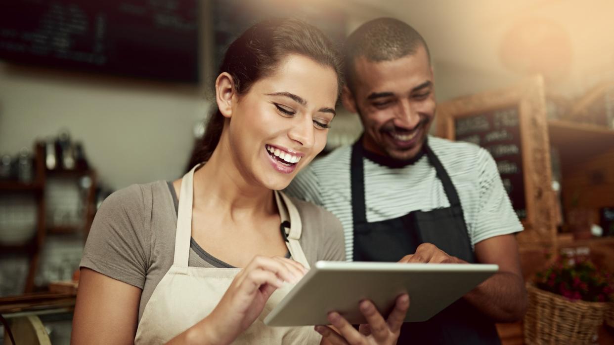 A female and a male cafe worker looking at a tablet computer smiling.