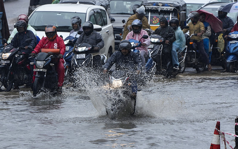 Motorists ride through a water logged street during rain. One motorcyclist rides past a row of traffic, with water splashing up in front of his wheels