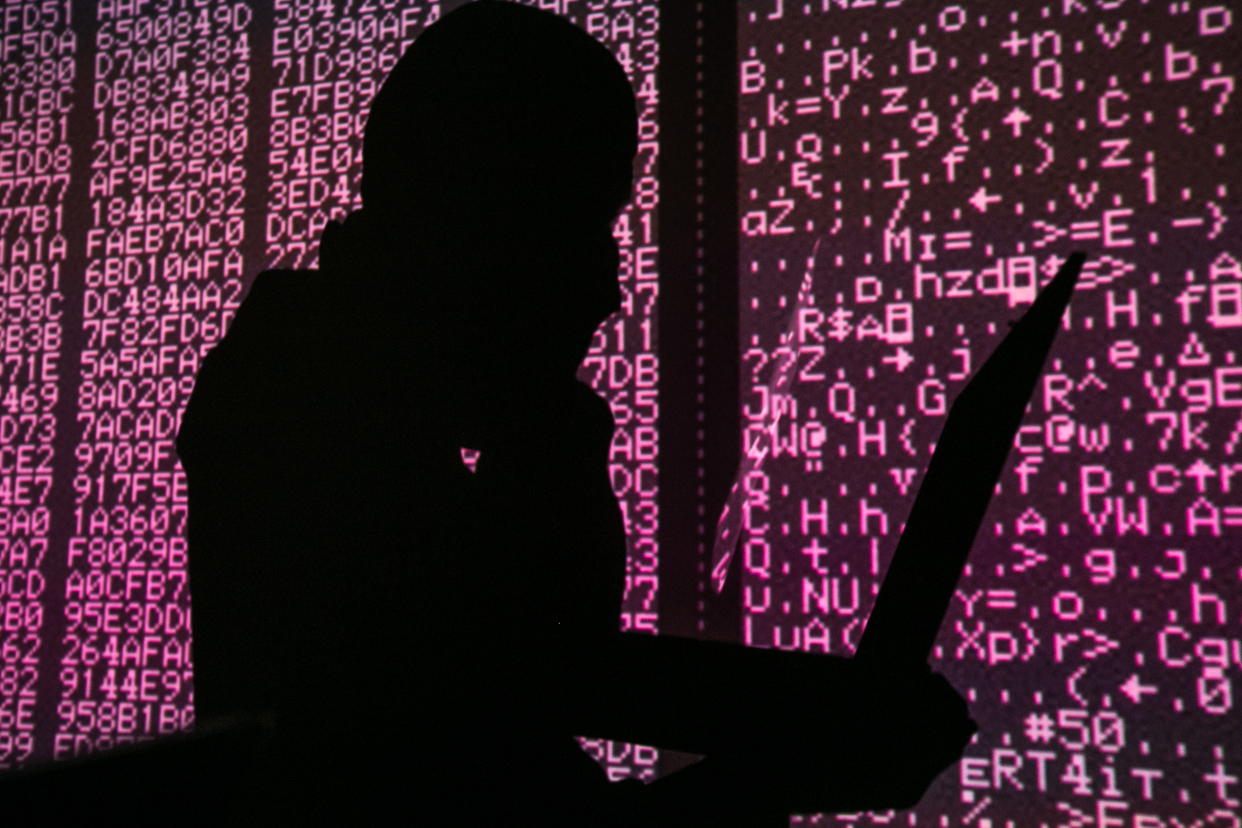 A silhouette of a man in a balaclava mask sitting at a laptop computer, with computer code in the background. (Sergei Konkov\TASS via Getty Images)