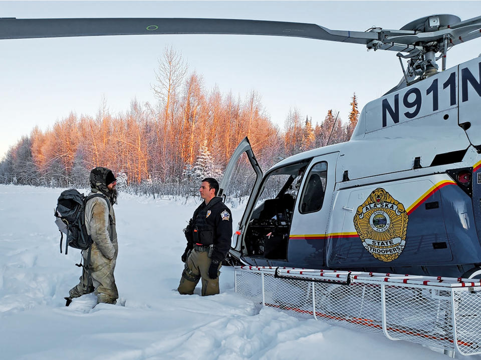 This Jan. 9, 2020 photo provided by Alaska State Troopers shows Tyson Steele, left, talking with Helo 3 Tactical Flight Officer Zac Johnson after Steele's rescue outside Susitna Valley, Alaska. Troopers rescued Tyson Steele, 30, who survived in a makeshift shelter after his remote cabin burned last month, after spotting him and an SOS sign in the snow. (Alaska State Troopers via AP)