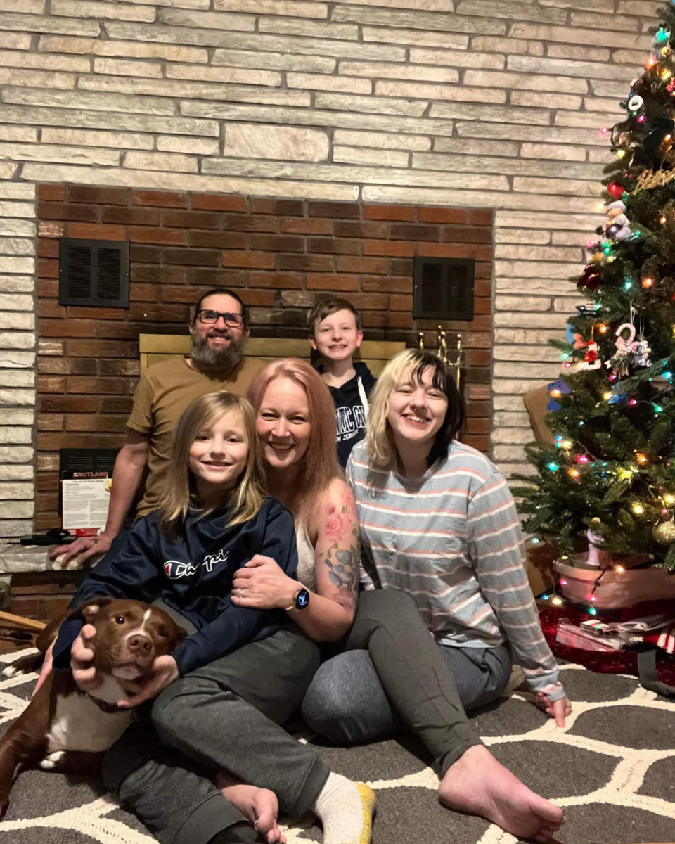 This Jan. 21, 2022 photo provided by Rebekah Hogan, center, shows her with her family next to their Christmas tree at their home in Latham, N.Y. More than a year after a bout with COVID-19, Hogan still suffers from severe brain fog, pain and fatigue that leave her unable to do her nursing job or handle household activities. “Is this permanent? Is this the new norm?’’ said the 41-year-old mother, whose three children and husband also have signs of the condition. “I want my life back.’’ (Courtesy Rebekah Hogan via AP)