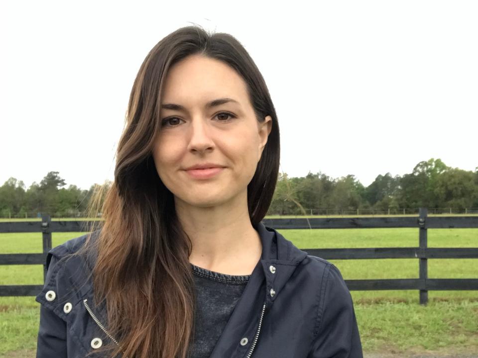 Portrait of a woman wearing a dark black jacket, backdropped by a green field and wooden fence