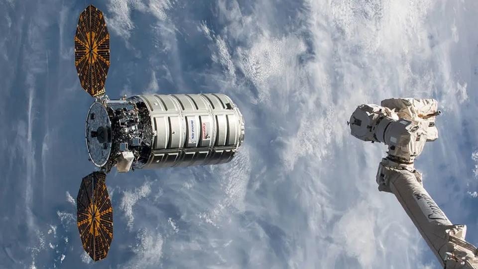 a spacecraft with two solar wings approaches a robotic arm in space. the earth and clouds are behind