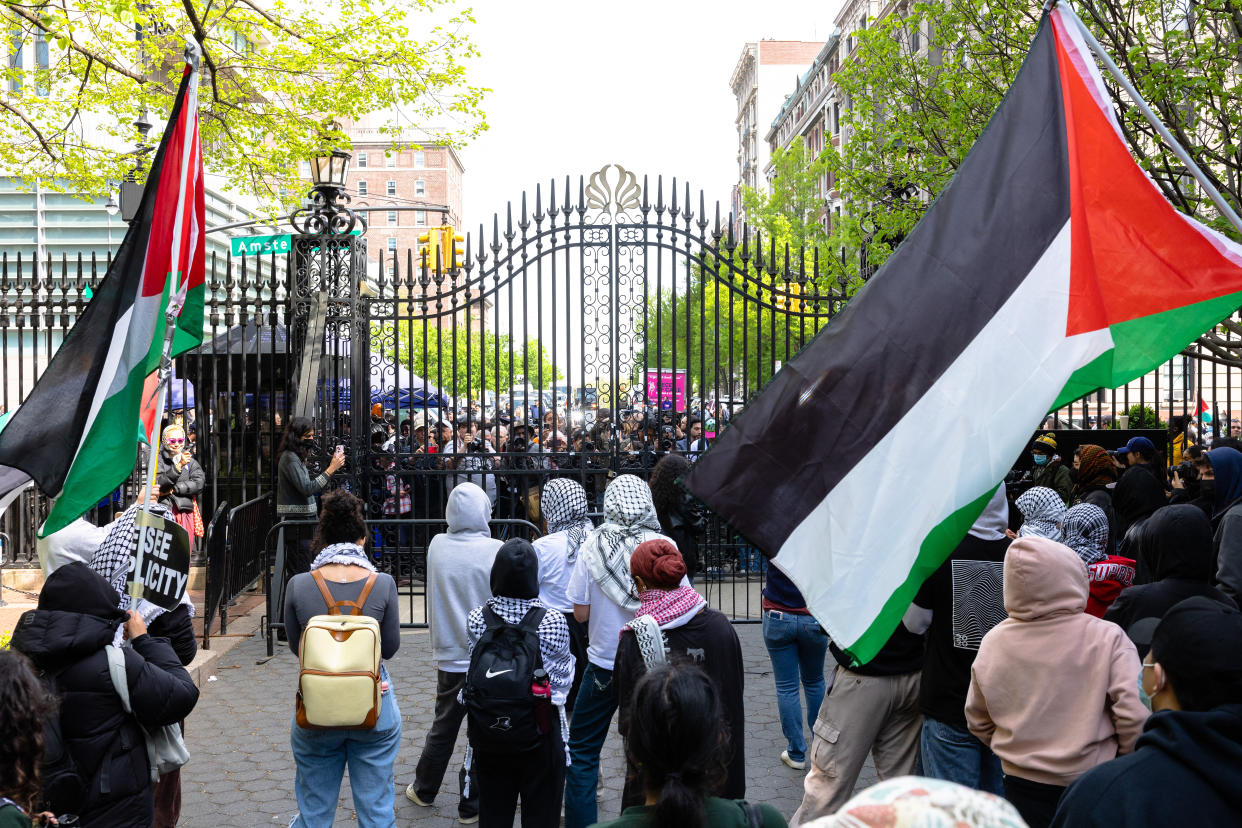 Dozens of people, some wearing headscarves and holding Palestinian flags, stand near an iron gate.