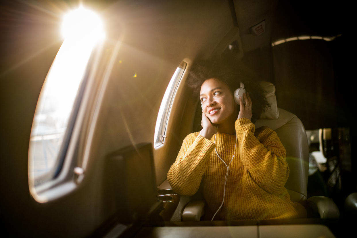 Young fashionable woman sitting on a private airplane and looking through a window while listening to music through headphones.