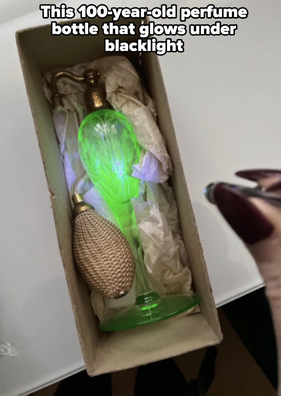 A vintage-style green perfume bottle with a bulb atomizer is placed in a box with crinkled paper. A hand with long, dark-colored nails is visible on the right