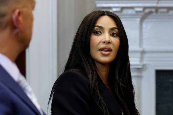 Kim Kardashian, wearing a black outfit, speaks during an event, with a partial view of a man's profile in the foreground