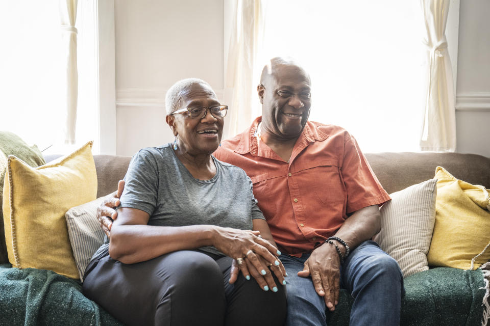 Elderly couple smiling, sitting closely on a sofa in a well-lit room, exuding warmth and companionship