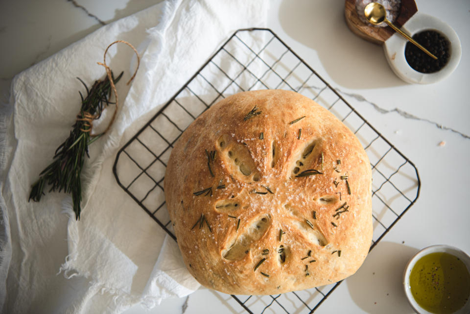 A freshly baked round loaf of bread on a cooling rack, with sprigs of rosemary on the side, a small bowl of oil, and a dish of ground black pepper