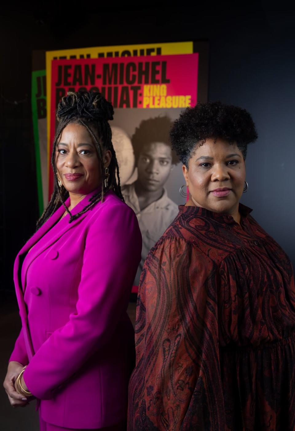 Two women stand back to back in front of an exhibition poster.