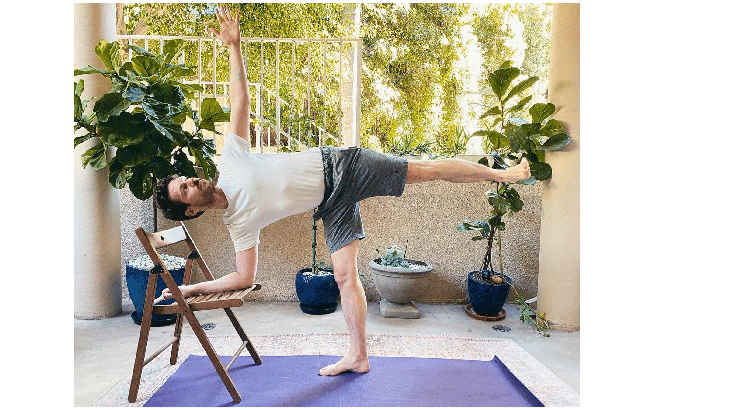 Man standing on a yoga mat practicing Half Moon Pose with his right forearm on the seat of a chair and his left leg lifted.