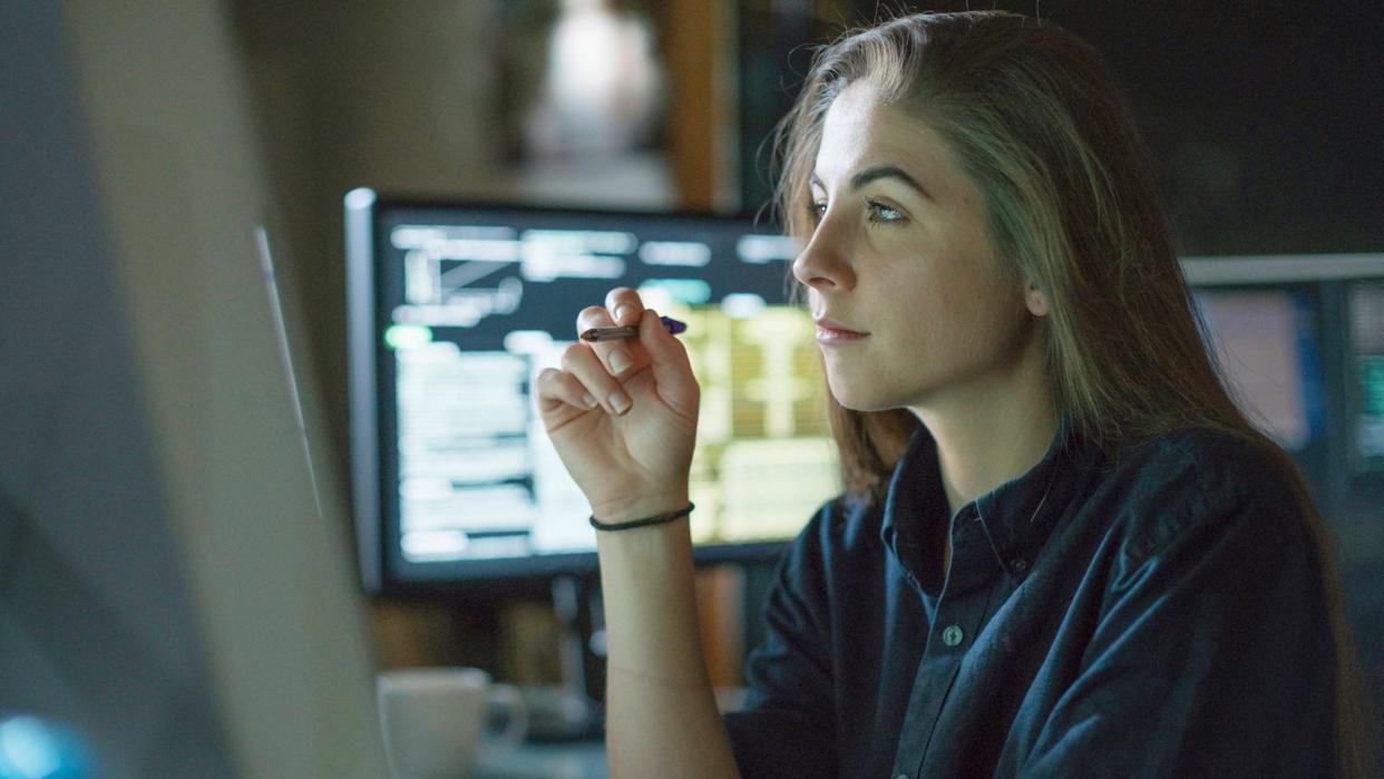 A young woman is seated at a desk surrounded by monitors displaying data, she is contemplating in this dark, moody office.