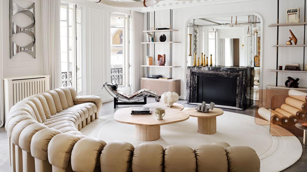 a white room, decorative molding, a trio of circular pendants, semicircle sofa in tufted beige leather, round rug, three cocktail tables, lounge chair, acrylic chair with cylindrical seat, black marble fireplace