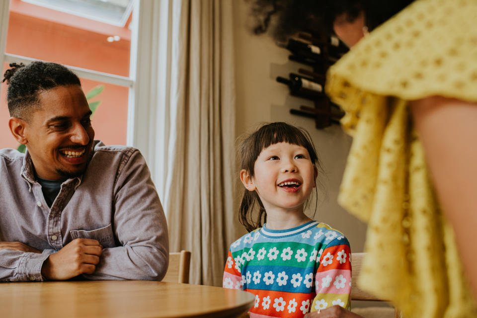 A cute young girl sits politely at a table. She looks up and smiles at a woman who is talking to her. A man in the background looks relaxed, and smiles as he watches over her.