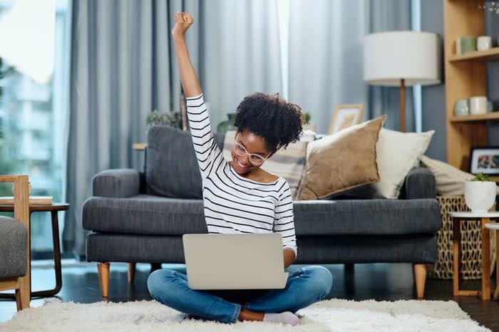 A woman sitting in front of her couch with her laptop and celebrating with her hand in the air.