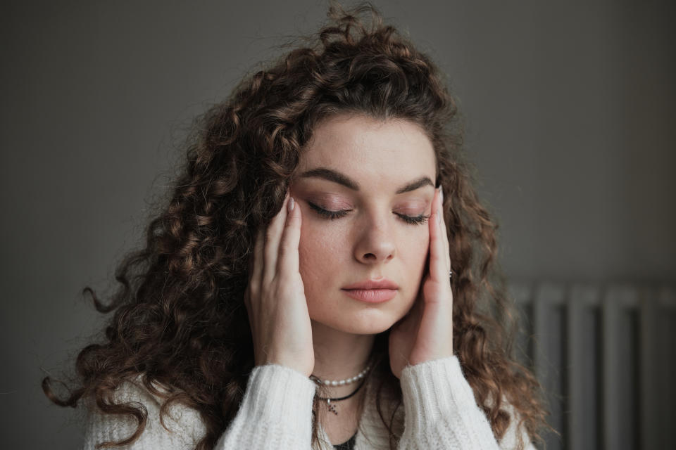 A woman with curly hair wearing a white sweater and pearl necklace stands with her eyes closed, gently touching her temples with both hands