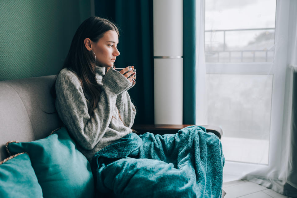 Shivering, pensive, frost brunette woman in sweater, wrapped in duvet sit on sofa and drink hot tea