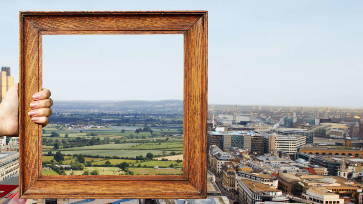 Young woman holding a picture frame. Outside the frame is a city, but inside the frame is a view of the countryside.