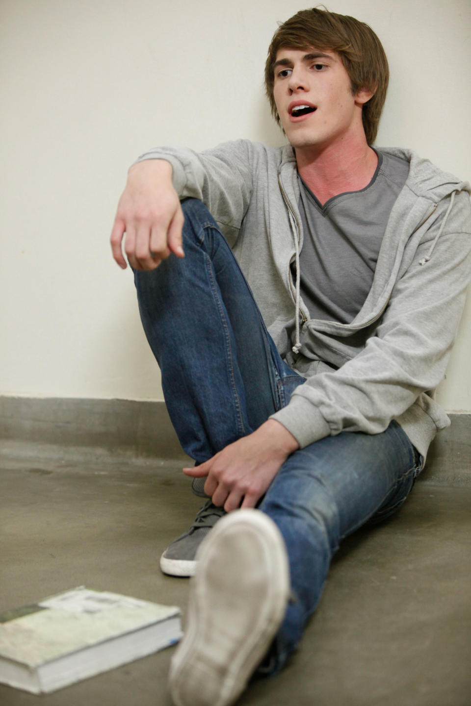 A young person is sitting on the floor, leaning against a wall, wearing a gray hoodie, gray shirt, and jeans. A book is on the floor nearby