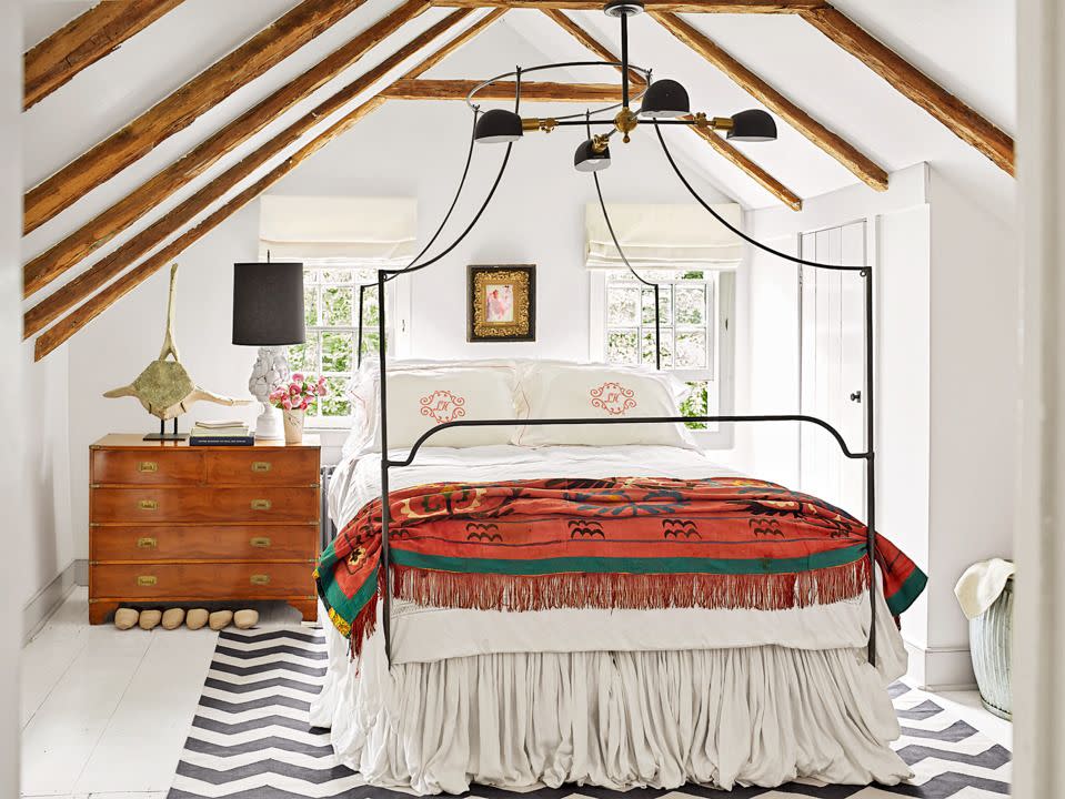 a wrought iron canopy bed complements the steep pitched ceiling in this upstairs bedroom with white walls and a wooden dresser and red blanket on the bed