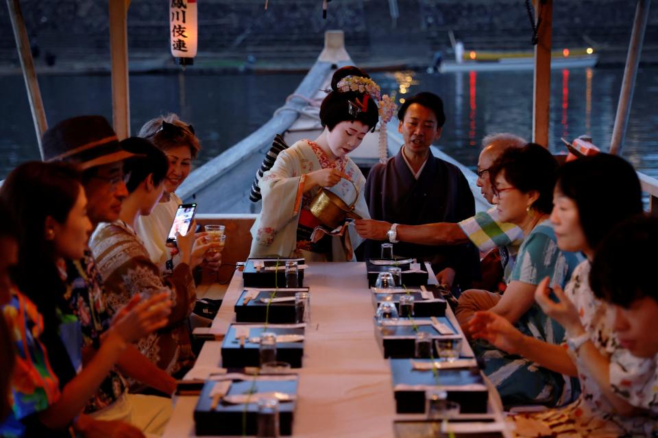 A group of people on a boat are served tea by a geisha in training.