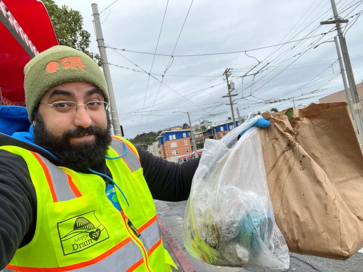 Bobak Esfandiari bearded man wearing glasses warm hat neon yellow visibility vest holding up a plastic bag and a paper bag full of garbage