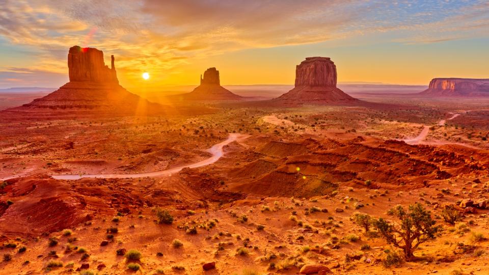 Monument Valley Navajo Tribal Park, with large rocky outcrops reaching up to the sunny sky. Small shrubs and trees are scattered across the Martian-like landscape.