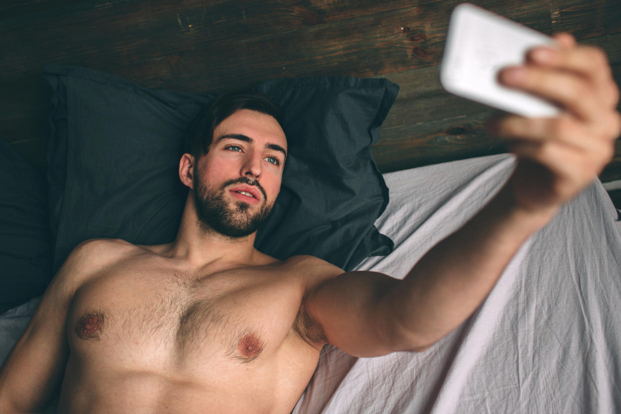 Naked bearded dark-hair handsome man shirtless in white bed taking a selfie.