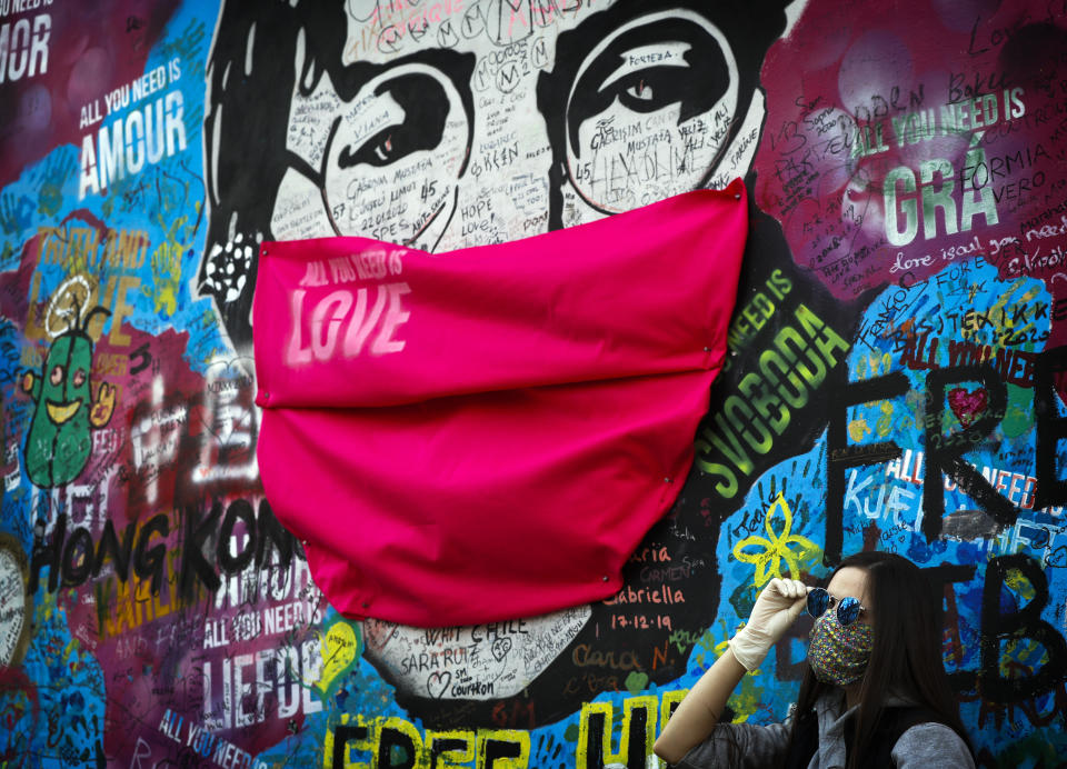 A woman poses for a photo by the "Lennon Wall" with a face mask attached to the image of John Lennon, in Prague, Czech Republic, Monday, April 6, 2020. (AP Photo/Petr David Josek)