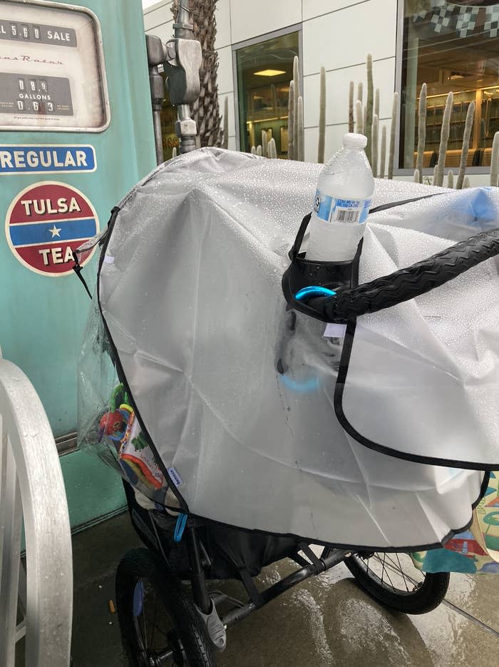 A covered stroller with a water bottle attached is parked next to an old gas pump labeled "Tulsa Tea." The area appears to be wet from rain