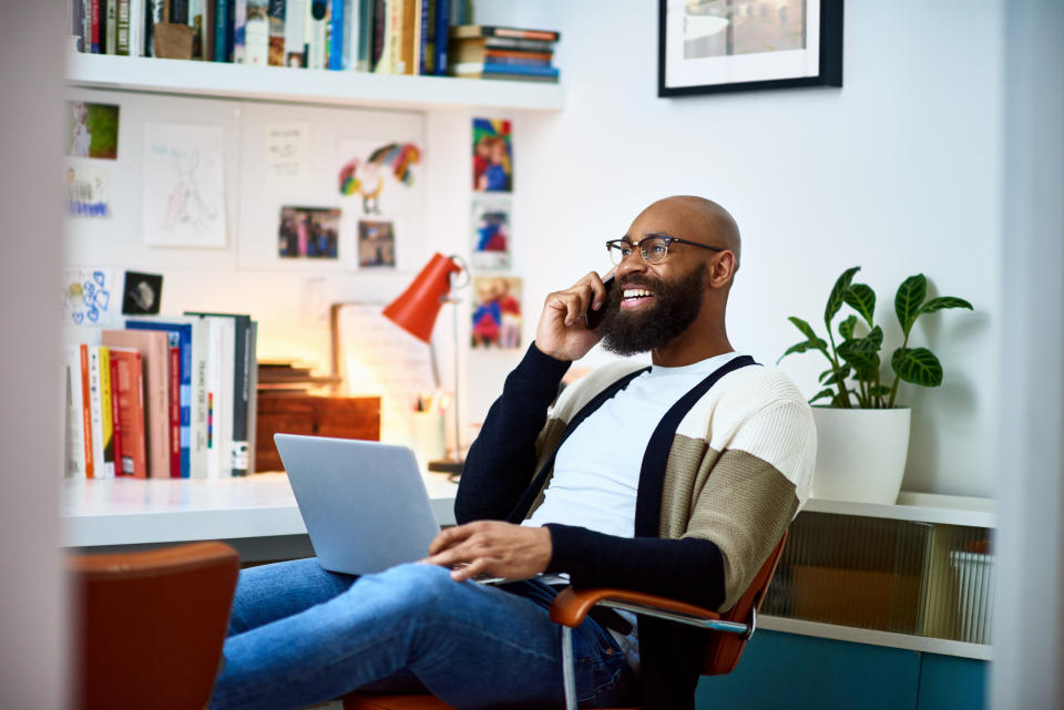 Man in a casual outfit sitting with a laptop, talking on the phone, bookshelf and plants in the background