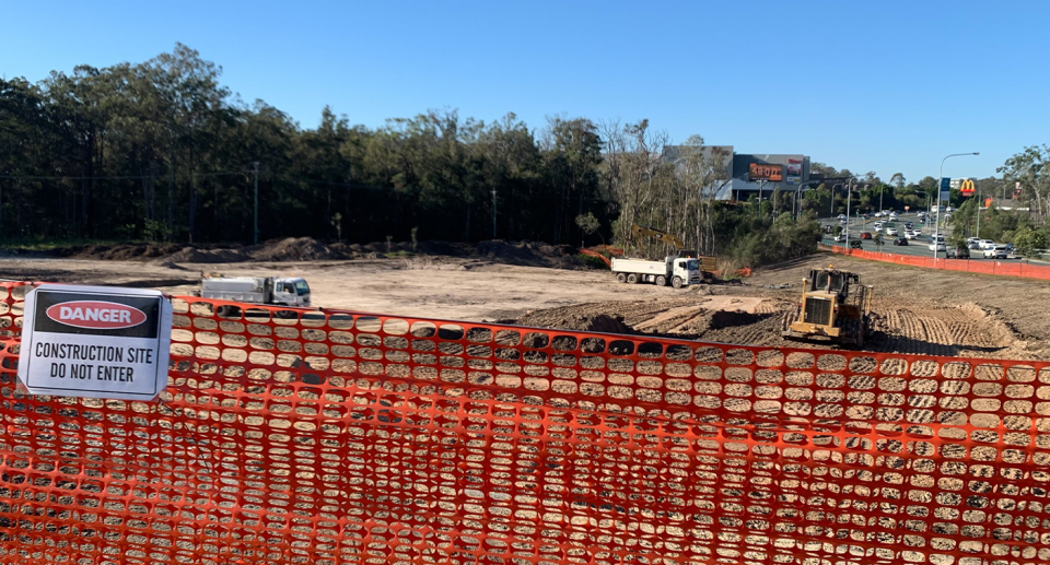 A construction site fence in front of clearing. McDonald's, KFC, Taco Bell can be seen in the background to the right and Westfield to the left.