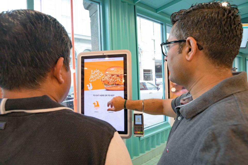 People try out the self-order kiosks at the re-opening of the Popeyes Louisiana Kitchen location on Canal Street in the French Quarter in New Orleans, Monday, March 28, 2022. The New Orleans restaurant is the first-ever Popeyes in the United States to feature modernized design including self-order kiosks, order ready boards, and dedicated areas for digital order pickup. Photo by Matthew Hinton