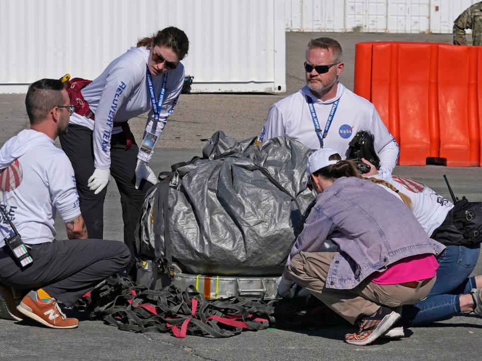 Several people in white shirts gather around a silver garbage bag containing a sample from NASA's OSIRIS-REx mission