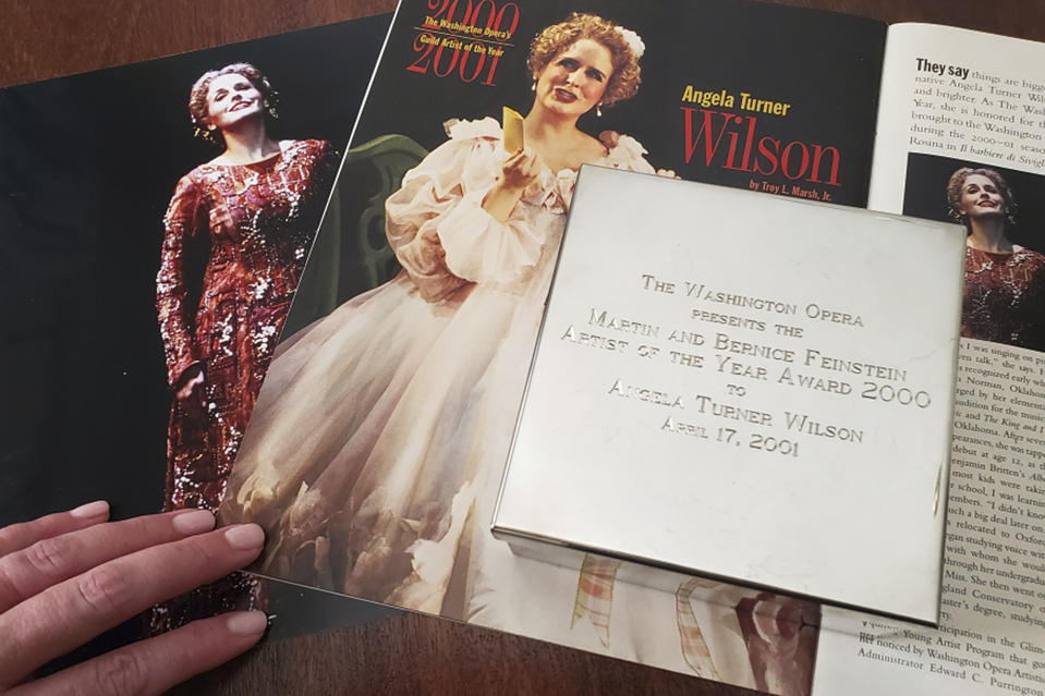 In this Tuesday, Sept. 2, 2019 photo provided by opera singer Angela Turner Wilson, she displays her 2000 Artist of the Year award from the Washington Opera next to a photo of herself from a 1999 performance of "Le Cid," left, and a magazine article in a Washington Opera magazine, at her home in Texas. An evening before a performance of "Le Cid," part of the Washington Opera's 1999-2000 season, she said she and Placido Domingo were having their makeup done together when he rose from his chair, stood behind her and put his hands on her shoulders. As she looked at him in the mirror, he suddenly slipped his hands under her bra straps, she said, then reached down into her robe and grabbed her bare breast. "It hurt," she told The Associated Press. "It was not gentle. He groped me hard." She said Domingo then turned and walked away, leaving her stunned and humiliated. (Courtesy Angela Turner Wilson via AP)