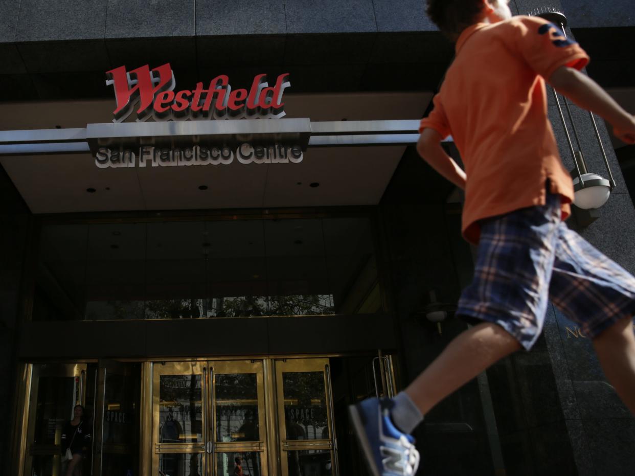 A young boy in an orange shirt and blue plaid shorts running past a set of doors at the entrance to the Westfield mall.