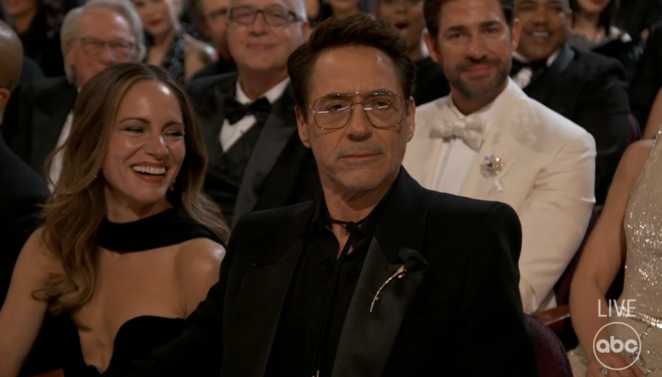 Audience at an event, man in tuxedo with glasses, woman smiling beside him, other attendees in background