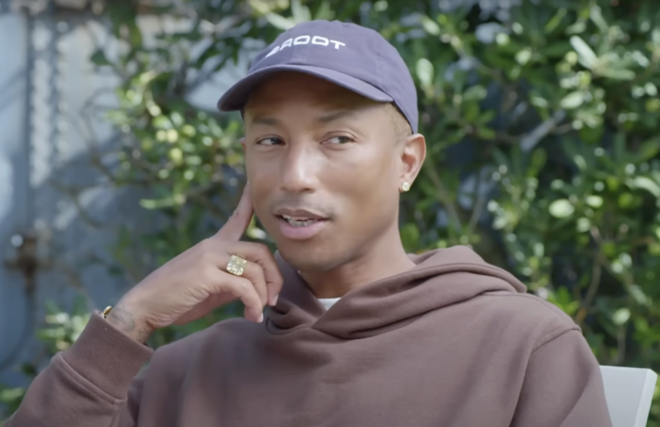 Pharrell Williams in a casual hoodie and cap, seated outdoors, smiling slightly with hand on chin