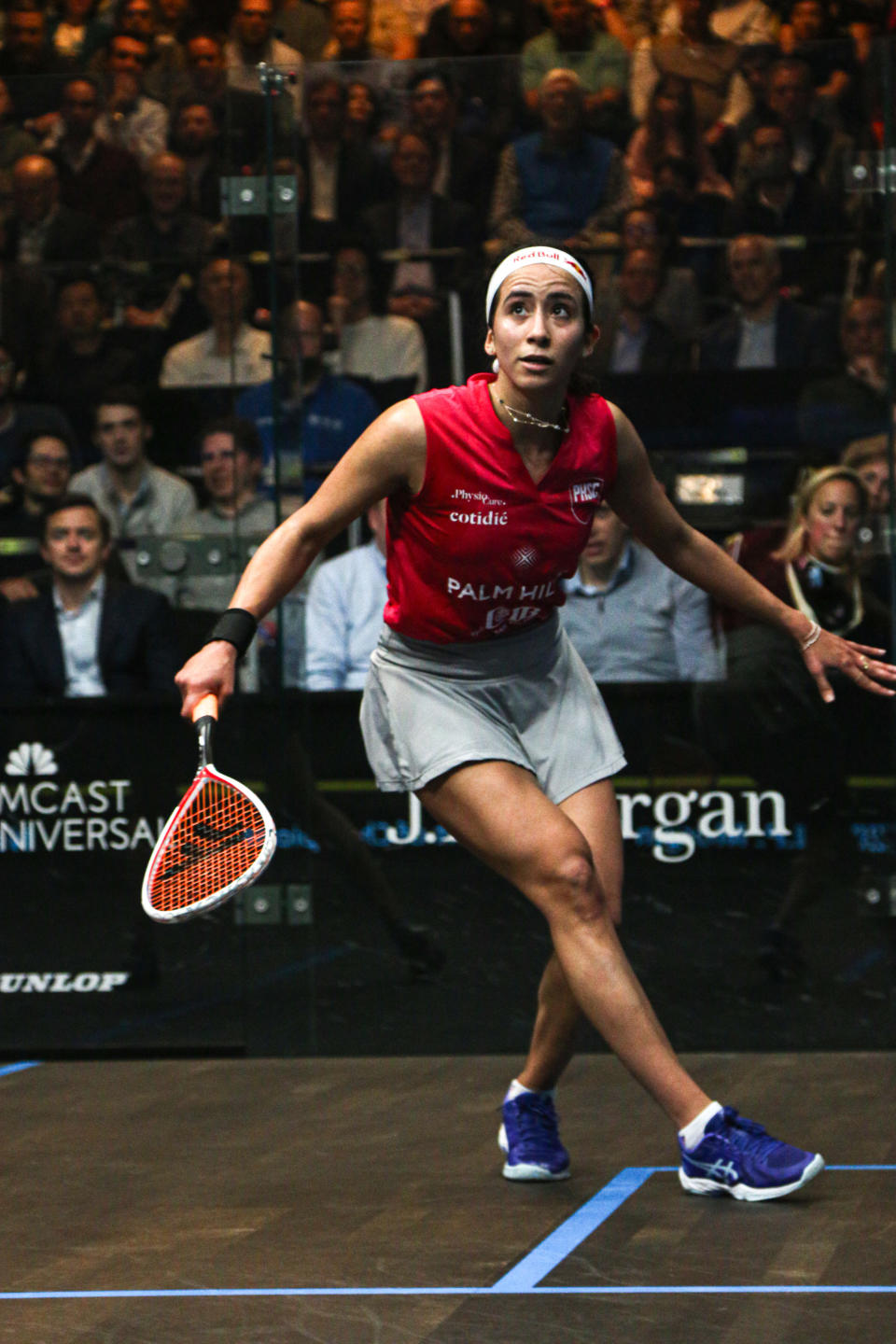 Raneem El Weleily, dressed in a sleeveless tracksuit, plays squash in front of an audience. She has a headband and is focused as she holds a squash racket