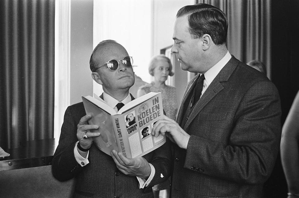 Un hombre bajito, calvo, con gafas de sol y traje sujeta un libro abierto mientras habla con otro hombre más alto.